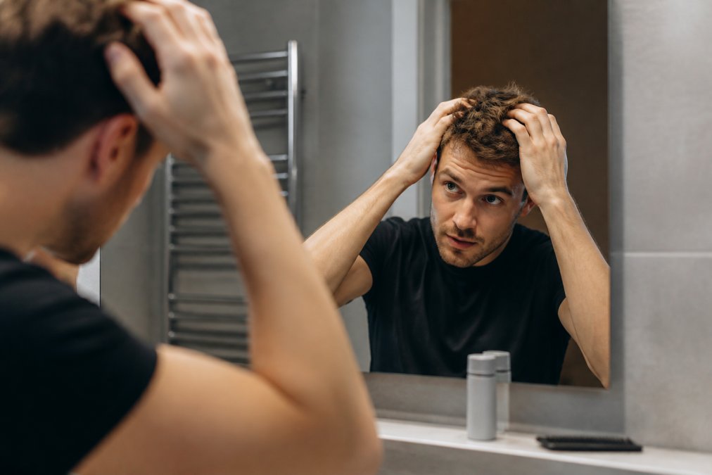 Man applying texturizing hair powder to his short hair in front of a bathroom mirror with a comb nearby