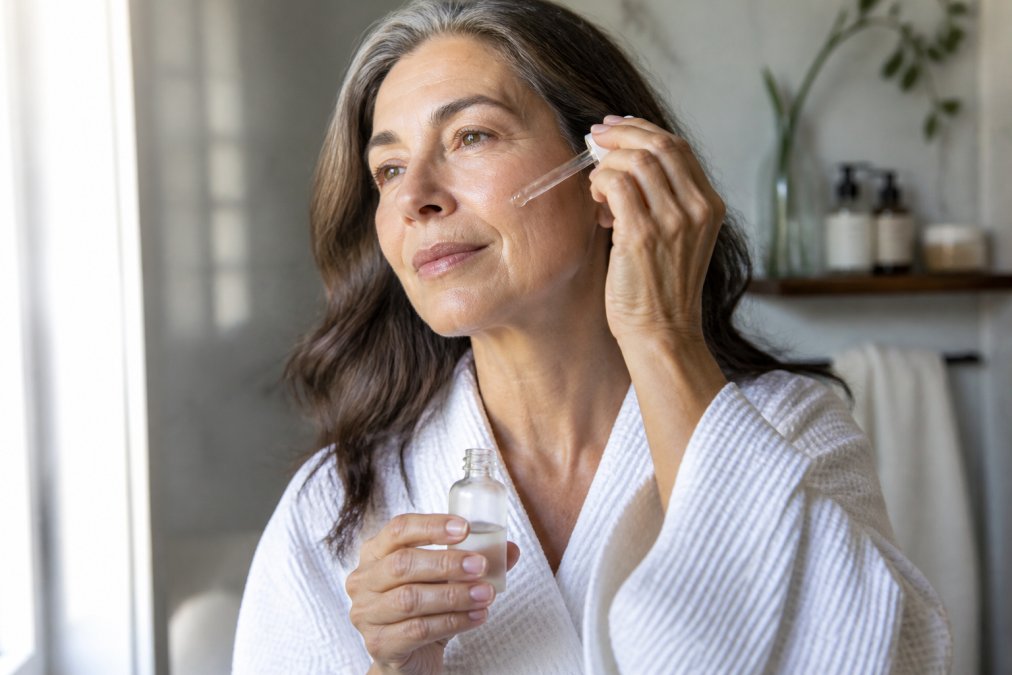 Woman in her 50s applying lightweight serum to cheek, natural morning light from window illuminating her face, visible glow on skin