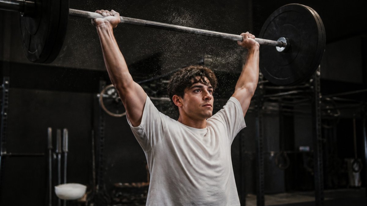 A lifter mid-overhead press in a plain, loose-fitting cotton t-shirt, fabric draping naturally at the shoulders and arms, shot in a dimly lit training gym with chalk dust in the air
