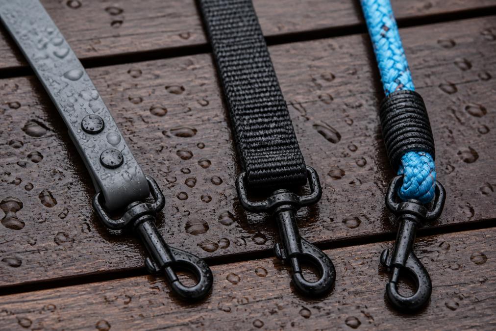Close-up of three different dog leashes laid side-by-side on a wet wooden deck: a biothane leash, a PVC-coated webbing leash, and a polypropylene rope leash. Water droplets visible on each surface to show water resistance.