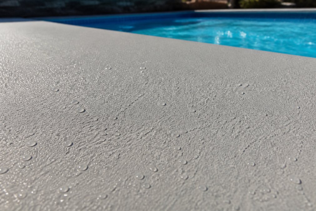 Close-up of freshly coated pool deck surface showing visible textured non-slip finish with water beads forming on the coating, barefoot-friendly appearance in natural sunlight