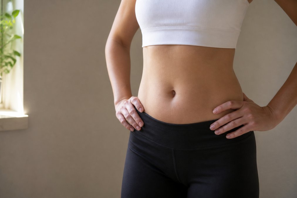 Close-up of a woman's midsection showing subtle body contouring improvements, photographed in soft natural window lighting against a neutral beige wall, her hands gently resting on her hips