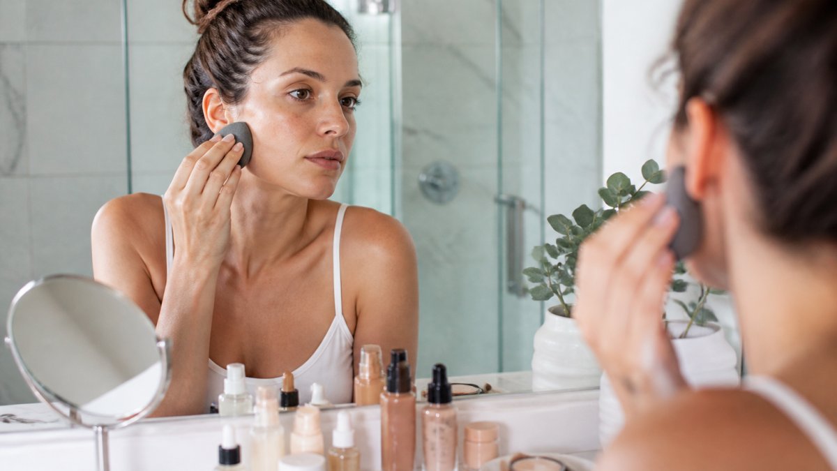 A woman with sensitive skin applying liquid foundation with a damp sponge at a bathroom vanity, surrounded by clean beauty products in glass bottles and a small mirror