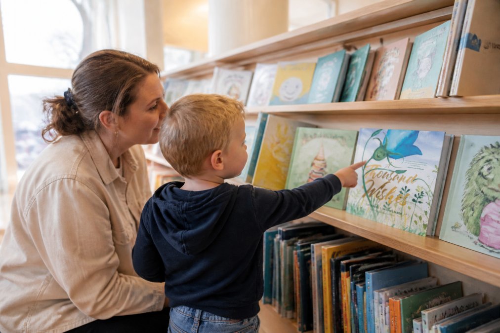 Forelder og barn som blar gjennom illustrerte barnebøker på bibliotekshylle, med fokus på fargerike omslag og barnet som peker på en bok som fanger oppmerksomheten, naturlig dagslys fra vindu