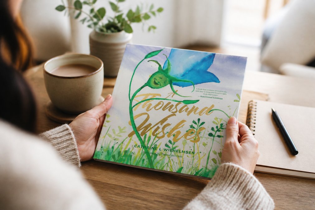 Nærbilde av hender som holder en vakker illustrert barnebok med detaljerte illustrasjoner, sittende ved et bord med en varm drikke og notepad i bakgrunnen, hjemmemiljø med naturlig dagslys