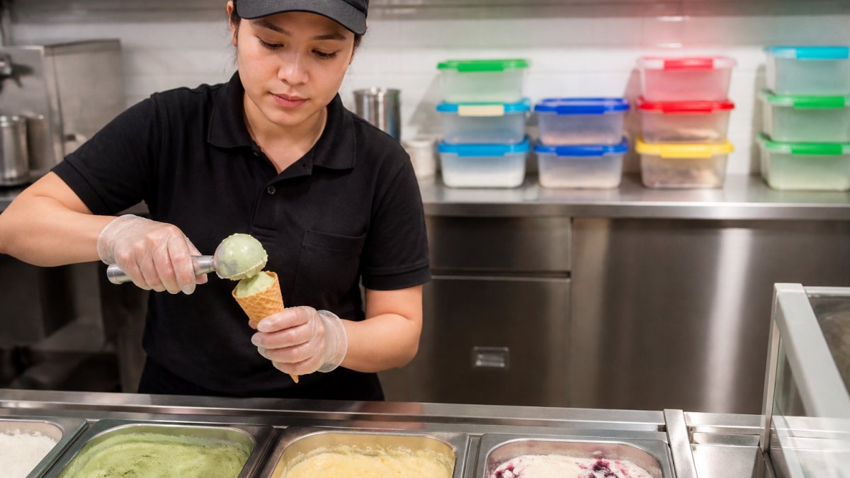 Professional café staff member carefully scooping nut-free ice cream into a cone using dedicated stainless-steel utensils, with clearly labelled containers and colour-coded storage containers visible in the background, bright café lighting