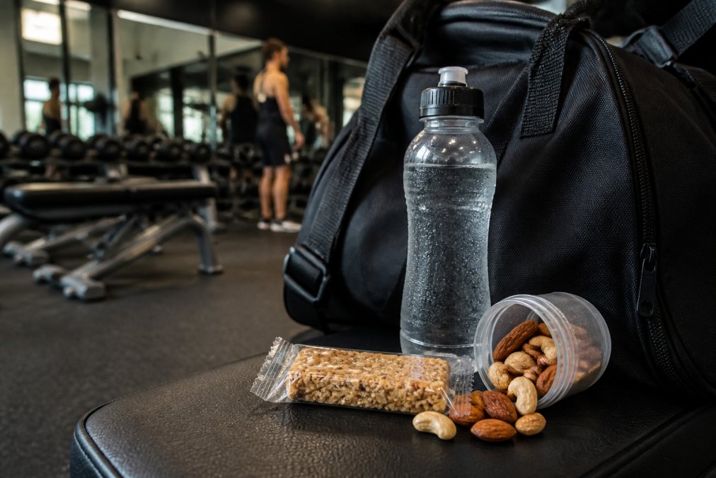 Gym bag on a bench with a water bottle and nutrient dense snacks like nuts and an energy bar ready for a workout.
