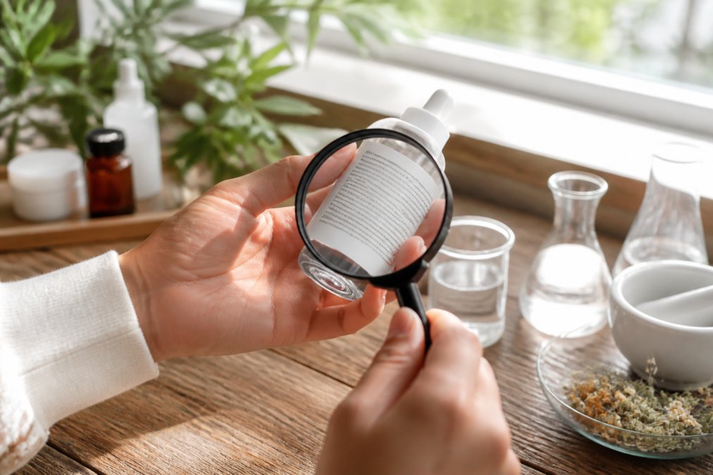Close-up of hands holding a glass skincare product bottle, examining the detailed ingredient list with a magnifying glass in natural daylight near a window