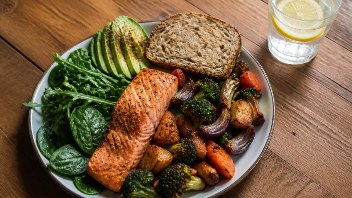 Colorful plate of whole foods including grilled salmon, roasted vegetables, leafy greens, and whole grain bread, with a glass of water and lemon, shot from above on a wooden table