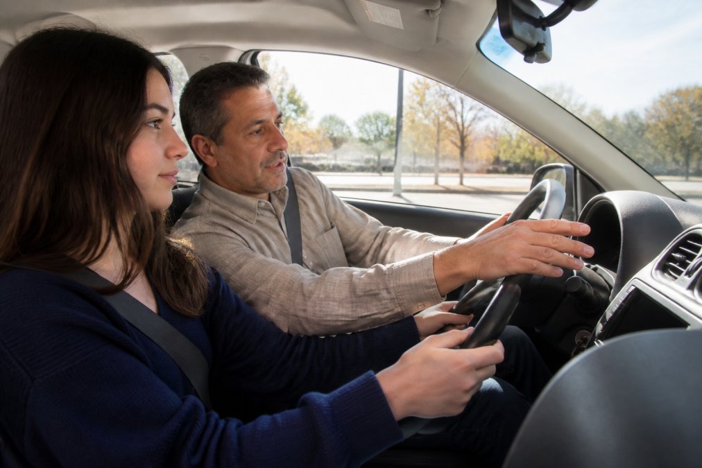 Experienced driving instructor providing calm, patient instruction to a nervous student inside a car during a lesson, with the instructor's hands positioned on the steering wheel demonstrating proper technique and the student focused on the road ahead