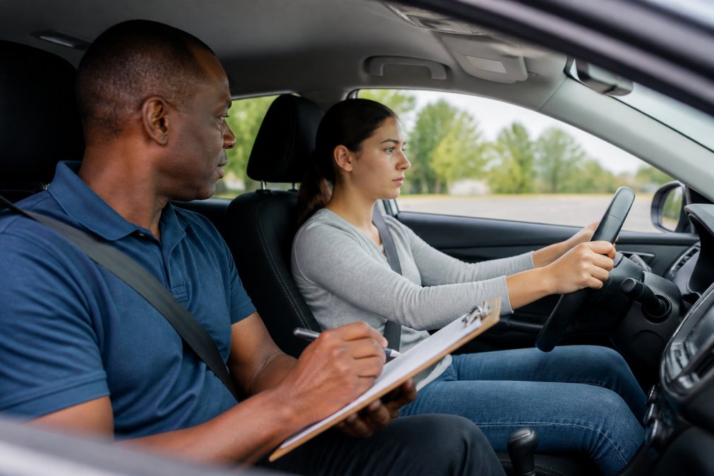 Nervous beginner driver gripping the steering wheel in a car, with an examiner in the passenger seat holding a clipboard, calm and professional atmosphere inside the vehicle