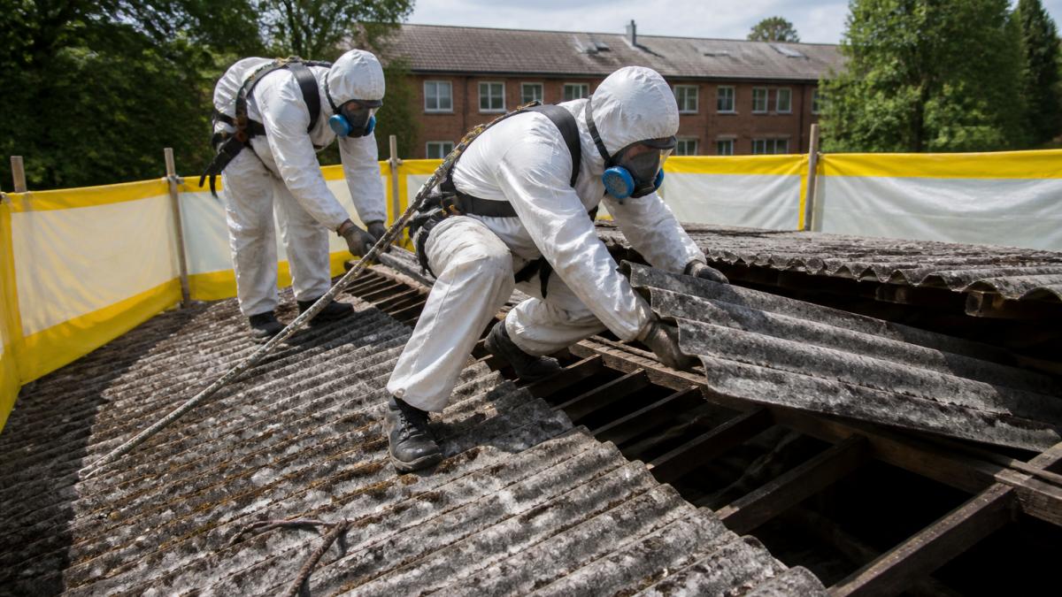 Certifikovaní pracovníci v bílých ochranných oděvech a respirátorech s modrými filtry provádějící demontáž azbestového materiálu ze střechy, s viditelným žlutým izolačním stanoveným kolem pracovní plochy a bezpečnostními pásy