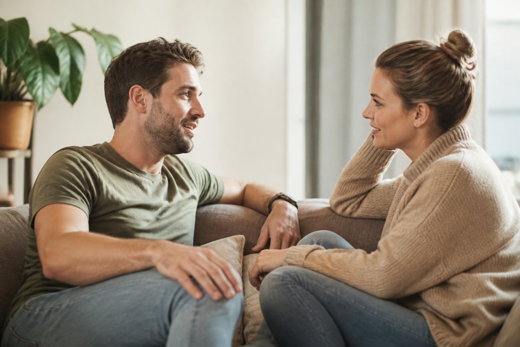 Een koppel zit tegenover elkaar op een comfortabele bank, beiden ontspannen en aandachtig naar elkaar luisterend, met oogcontact en open lichaamstaal
