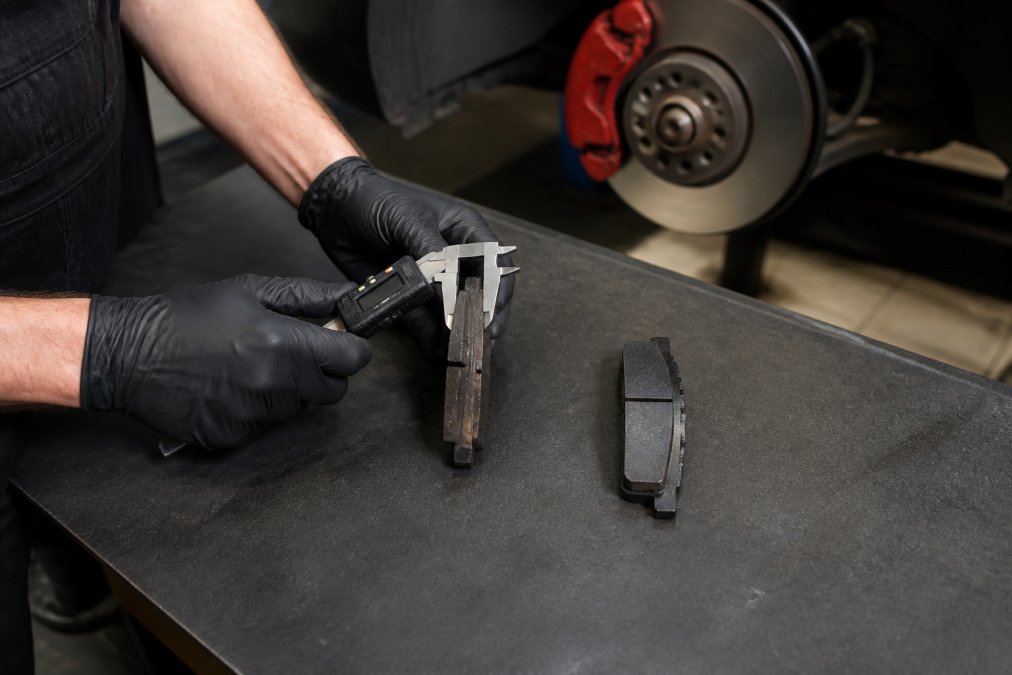 Certified technician measuring brake pad thickness with digital caliper tool, displaying both worn and new OEM brake pads side by side on workbench with measurement markings visible