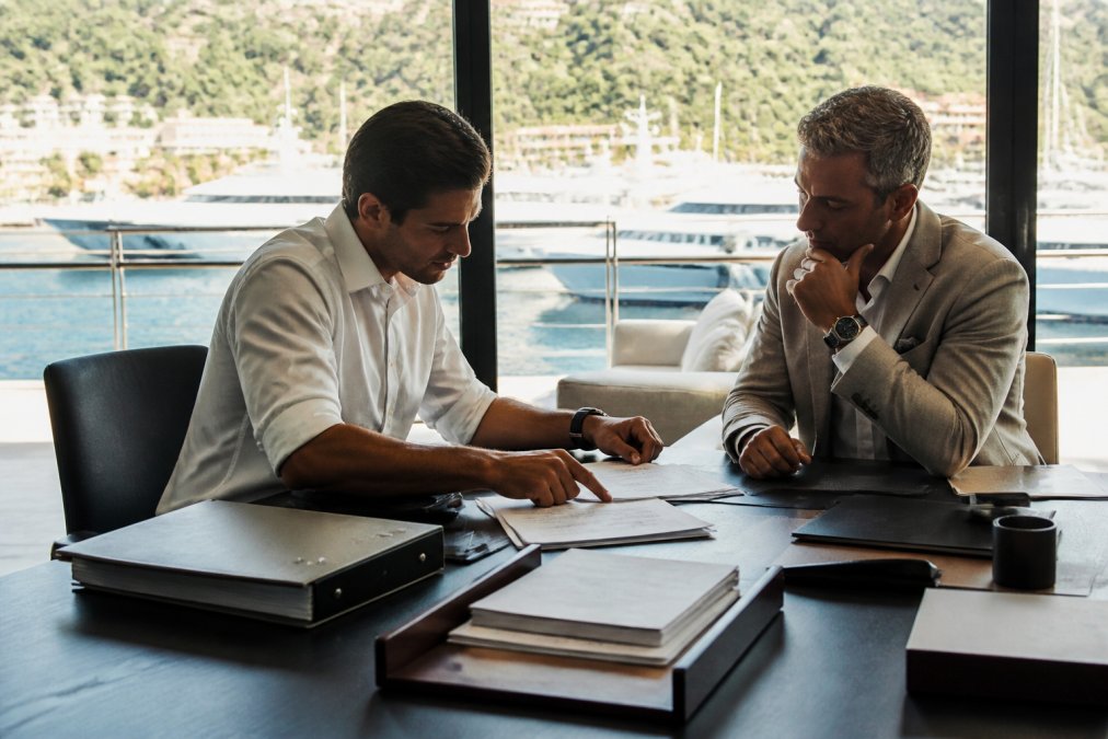 Professional yacht broker meeting with client in private office with secured documents and confidential files visible on desk, soft natural lighting from floor-to-ceiling windows overlooking marina
