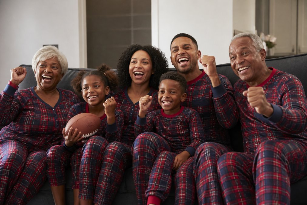 Multi-generational family wearing coordinated Team USA plaid onesies and matching loungewear, sitting together on a couch during a game day, smiling and showing team pride