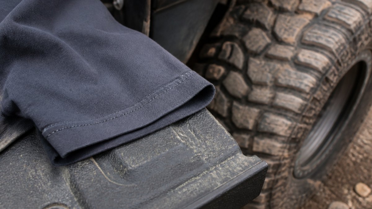 Close-up of a heavyweight cotton t-shirt sleeve showing the double-stitched hem, laid on a tailgate next to a muddy off-road tire