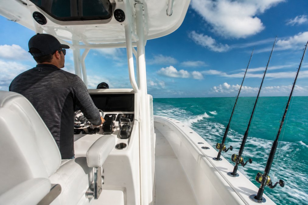 Captain standing at the helm of a center console boat navigating through blue-green offshore waters, with clear visibility of the navigation electronics and fishing rod holders mounted on the gunwales