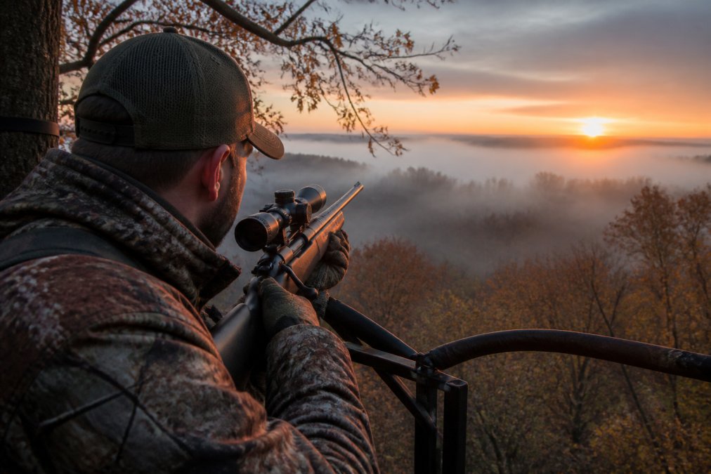 Hunter in a tree stand at dawn with rifle, surrounded by autumn woods and fog rolling through the valley
