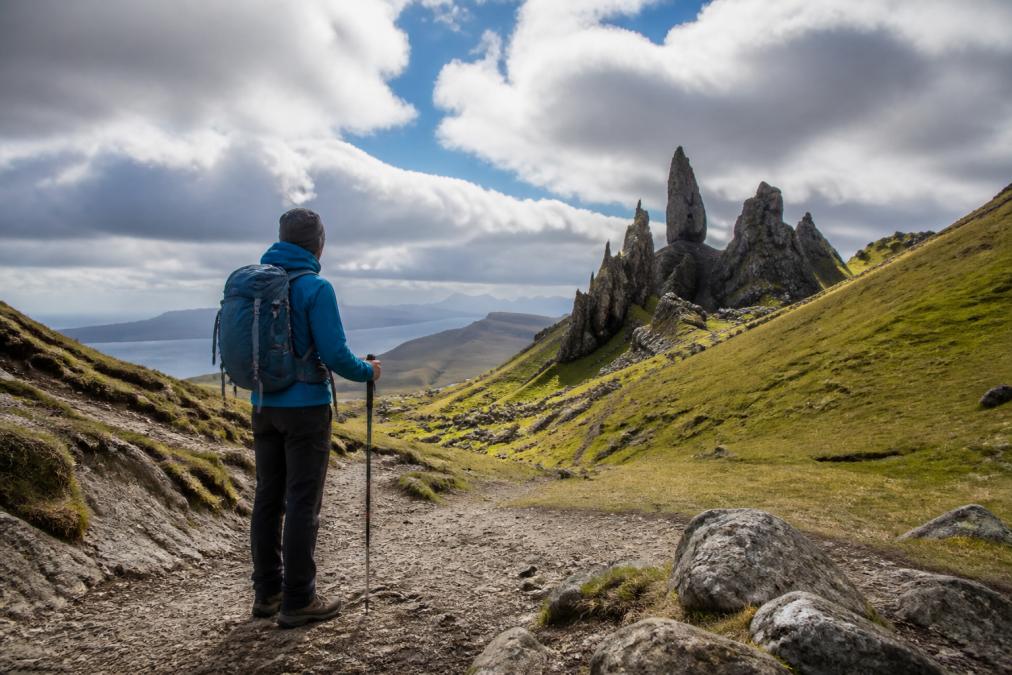 Hiker standing at the trailhead of the Old Man of Storr with the iconic rock pinnacle visible in the background under a dramatic Skye sky, wearing waterproof jacket and hiking boots