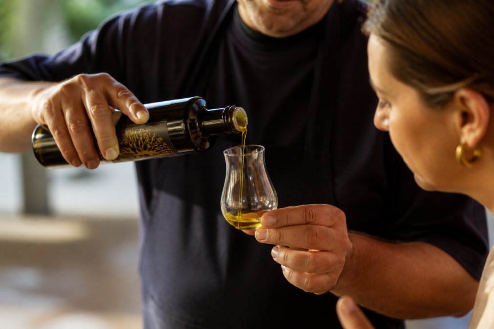 Close-up van handen die olijfolie in een glas gieten met zonlicht dat de kleur en textuur toont, tegen een neutrale achtergrond