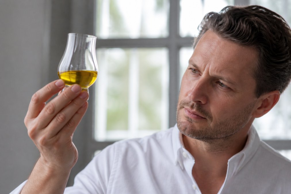 Close-up van een glazen proefglas met gouden olijfolie tegen een witte achtergrond, met een hand die het glas schuin houdt, helder natuurlijk licht