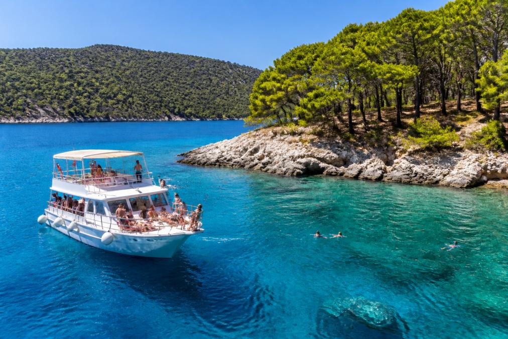 Güneşli bir yazın günü, turkuaz renkli Ölüdeniz denizinde beyaz tekne, yolcuların güneş altında eğlendiği, berrak su, kıyıdaki yeşil çam ağaçları ve kayalık sahne