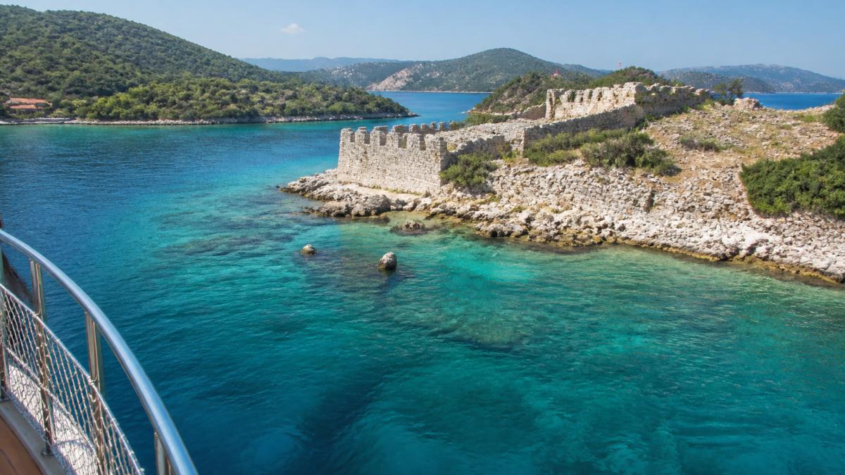 Tekne güvertesinden çekilmiş fotoğraf, turkuaz sular içinde Şövalye Adası'nın tarihi kale kalıntıları görülüyor, arka planda yeşil tepeler ve diğer küçük koylara genel bakış