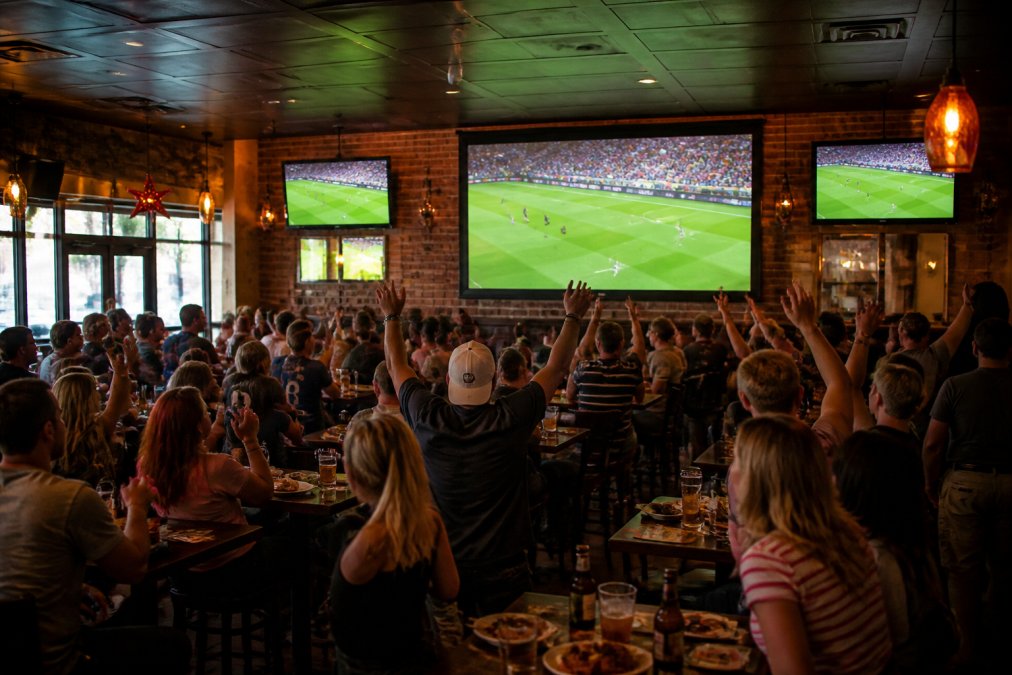 Wide shot of a lively sports bar interior with multiple HD TVs showing a soccer match, patrons cheering at tables, and warm ambient lighting