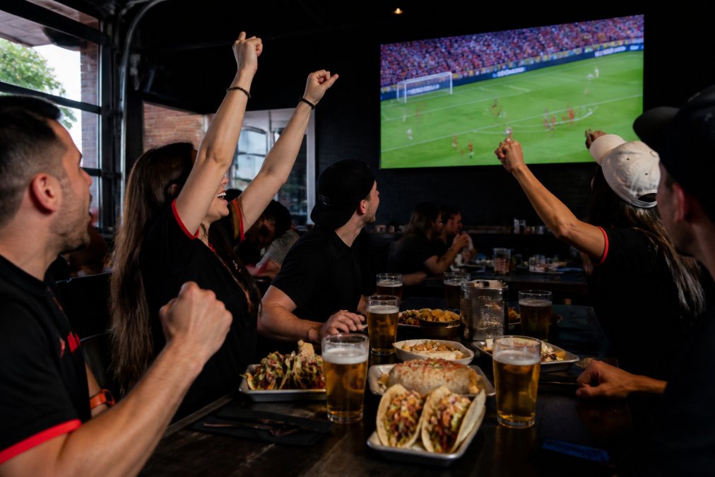 A lively group of friends cheering at a bar table, eyes fixed on a large-screen television showing a live soccer match, with plates of tacos and burritos and frosty draft beers on the table