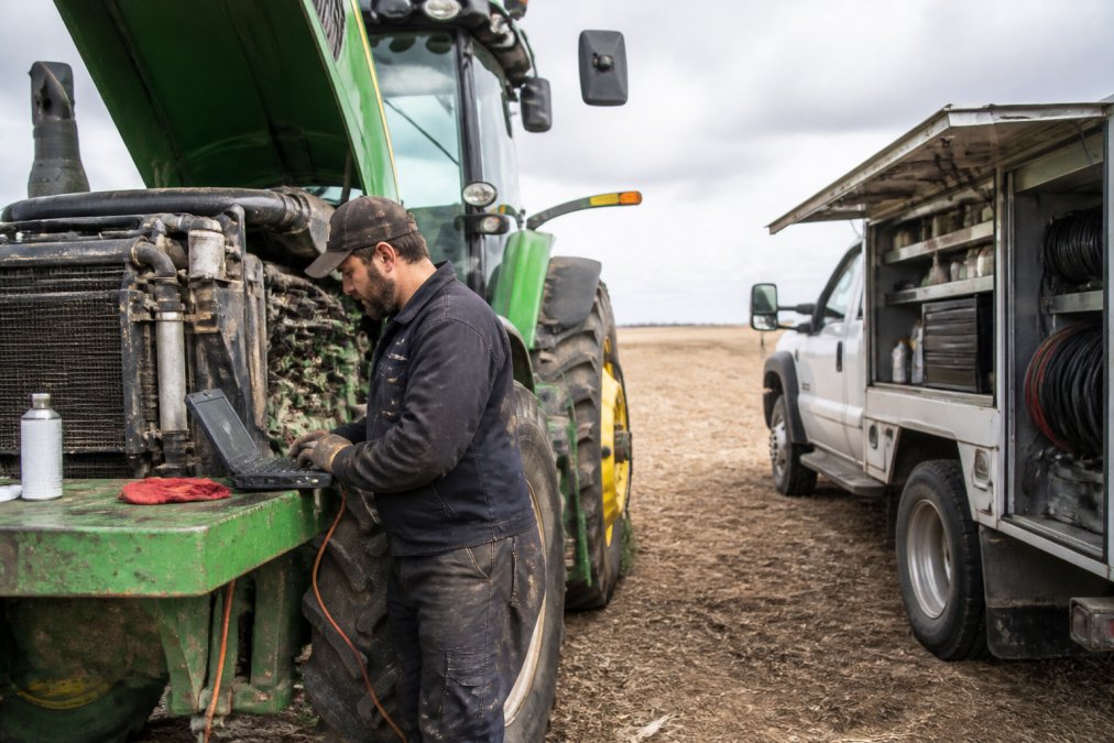 A mobile technician in work gear using a diagnostic laptop connected to a large farm tractor engine in an open field, service truck parked nearby with open equipment bay revealing organized tools and parts