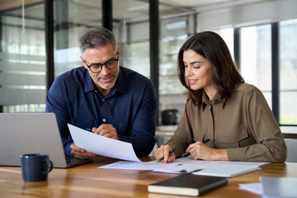 L&D manager and new corporate trainer sitting together at a desk reviewing printed documents and a laptop, in a professional office setting with warm natural light coming through a large window