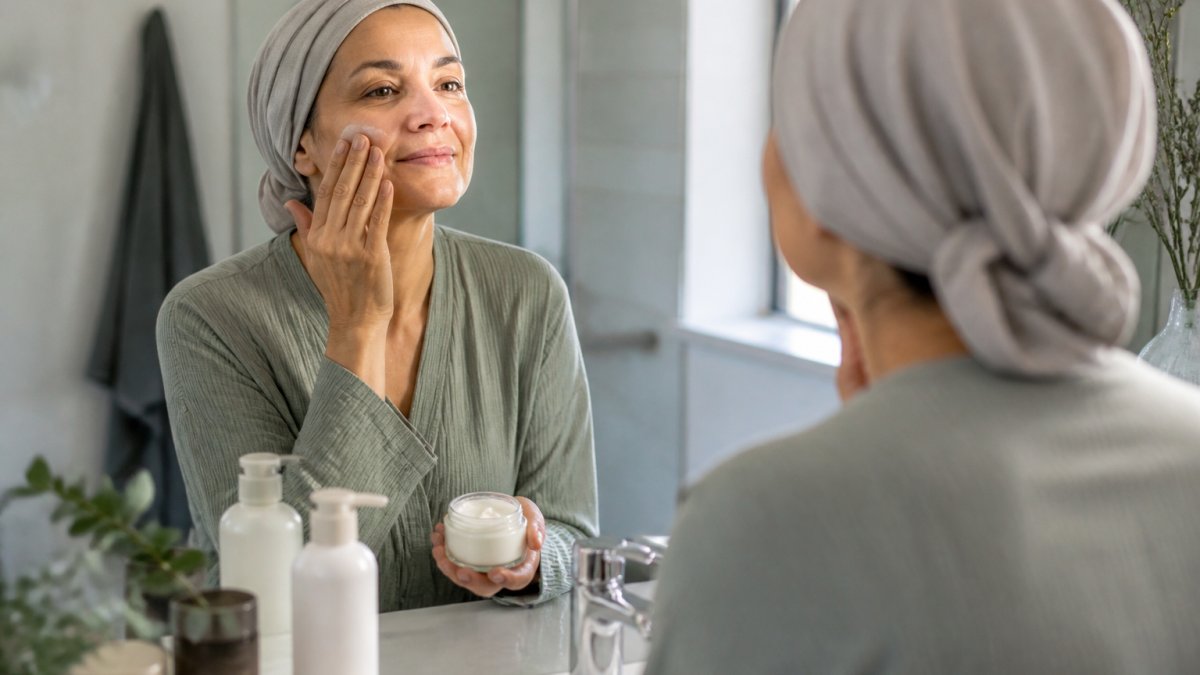 A woman with a calm, gentle expression applying moisturizer to her face in a softly lit bathroom, wearing comfortable clothing, with a few simple unbranded skincare products on the counter