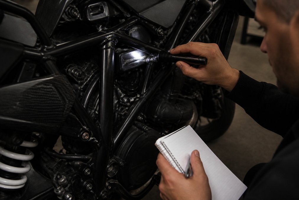 Close-up van iemands handen die het VIN-nummer op een motorfiets-frame controleren met een notitieblok en zaklamp in een werkplaats