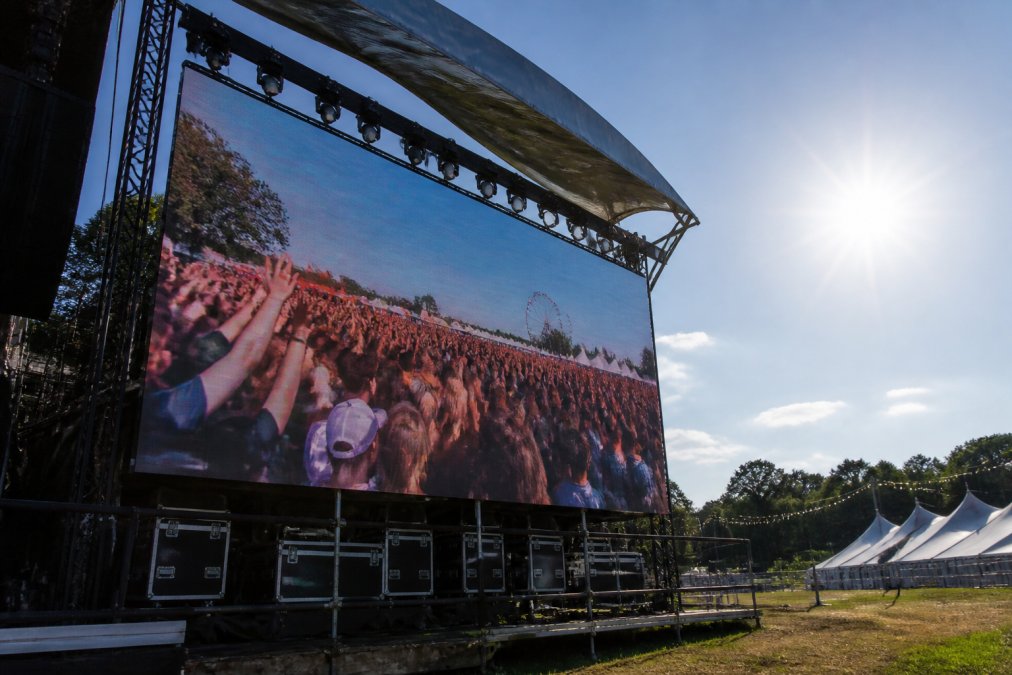 Large outdoor LED screen displaying vibrant colorful content at an outdoor festival under bright sunlight, with visible weather protection canopy overhead and technical equipment mounted securely at the base