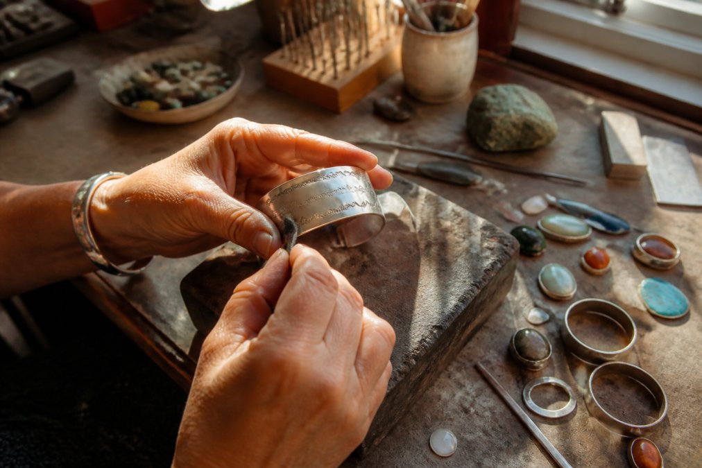 Artisan shaping a silver cuff bracelet on a workbench to create unique handmade jewelry gifts in a sunlit studio.