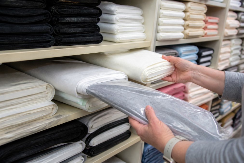 Close-up of hands examining and selecting various interfacing bolts and packages on a retail shelf, showing different weights, colors, and brands of fusible and sew-in materials in natural lighting