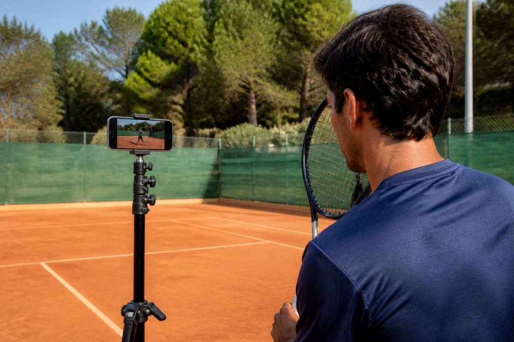 Adult tennis player on court holding a smartphone mounted on a tripod, analyzing their forehand stroke in real-time with app feedback displayed on screen, natural daylight on clay court