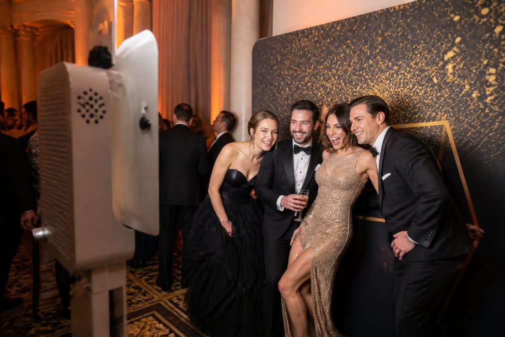 Elegantly dressed gala guests laughing and posing together in front of an open air photo booth with custom gold and black backdrop, with soft uplighting illuminating the setup and surrounding guests mingling in the background
