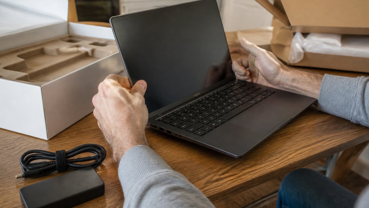Close-up of hands inspecting an open-box laptop on a kitchen table, checking the screen, ports, and charging cable next to the original box and packing materials