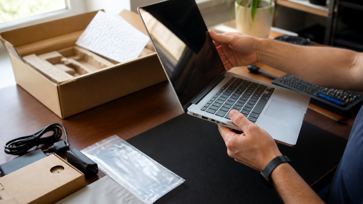 Close-up of hands inspecting a laptop screen and ports at a desk, with the original box and accessories laid out nearby, natural window light