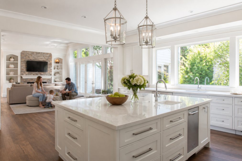 Bright modern kitchen with white cabinetry and large windows flooding the open concept space with natural daylight, family members visible in adjacent living area