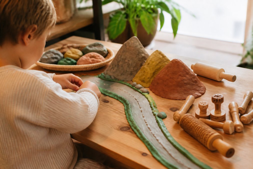 Een peuter die met natuurlijke speelklei in bouwkleuren een weg en bergen vormt op een houten tafel, met houten stempels en rollers ernaast, warm daglicht, huiselijke omgeving