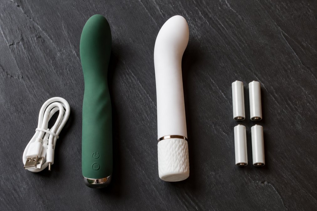 Een donkergroene oplaadbare vibrator en een matwitte batterijvibrator naast elkaar op een zwarte leistenen ondergrond, met een opgerolde USB-oplaadkabel en vier losse alkalinebatterijen ernaast, zacht zijlicht