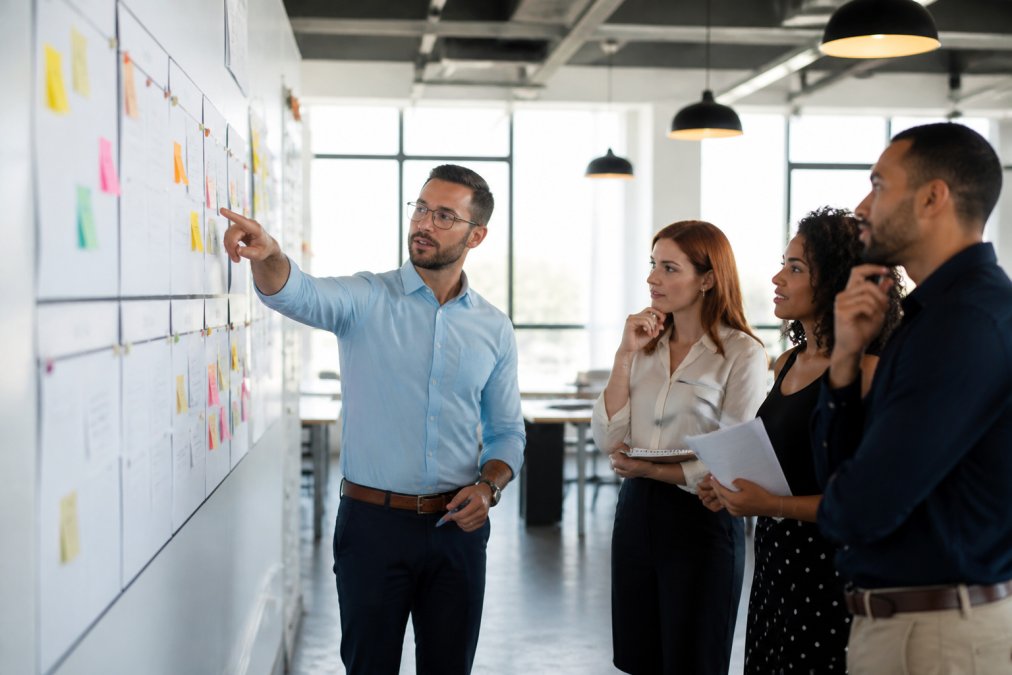 A sales team gathered around a large whiteboard covered in account lists, sticky notes, and tier labels, actively discussing strategy in a modern open-plan office with large windows and bright natural light