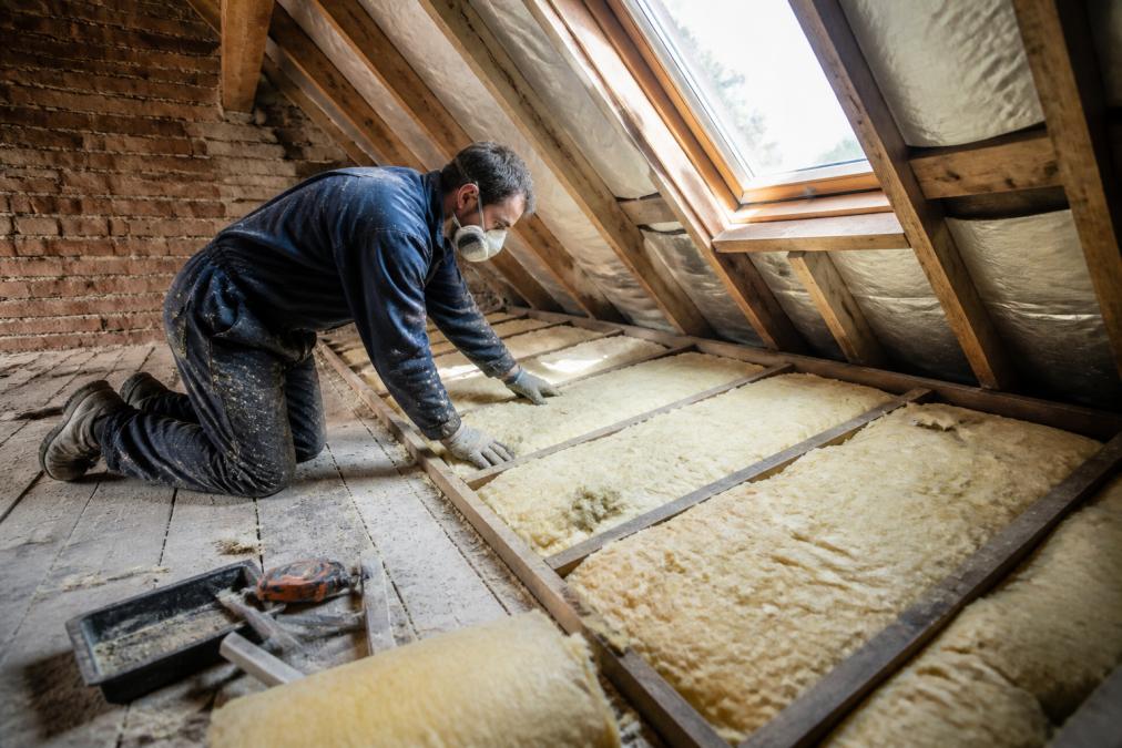 Un artisan en combinaison isolant les combles d'une maison ancienne en briques, pose de laine minérale entre les chevrons, lumière naturelle par une lucarne, poussière et outils sur le plancher