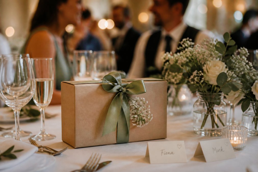 Close-up of a personalised cocktail box on a wedding reception table next to handwritten place cards and a small floral arrangement, with guests chatting softly out of focus in the background