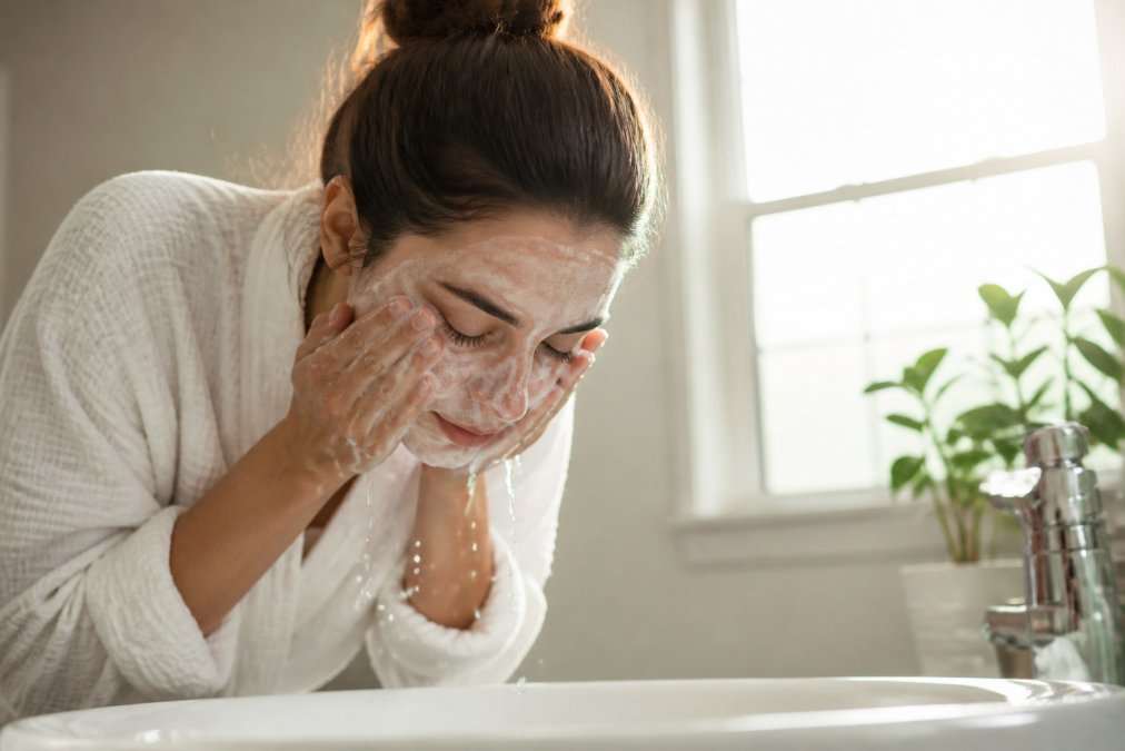 Woman's hands gently massaging a facial cleanser into her face over a white sink with soft morning light streaming through a window