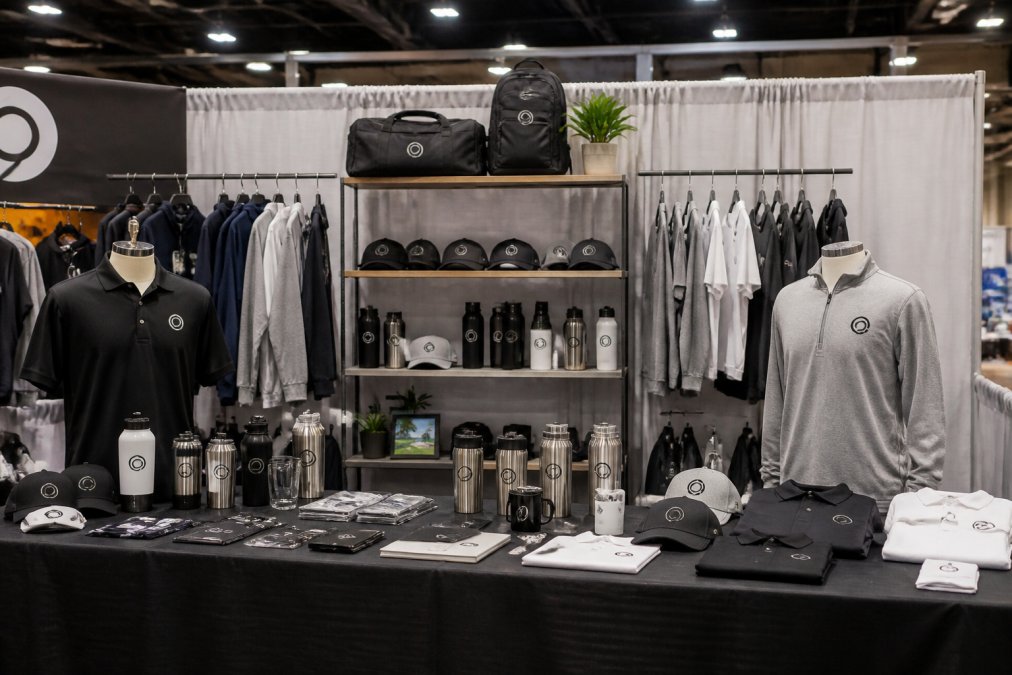 Trade show booth displaying branded merchandise including apparel, drinkware, and promotional items with company logos visible on display tables and shelves