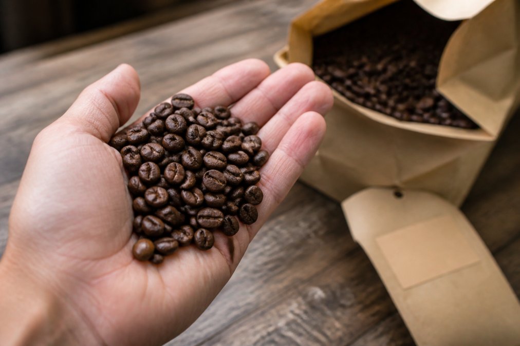 Close-up of glossy, richly colored whole coffee beans in an open palm, with a visible roast date label in the soft-focused background
